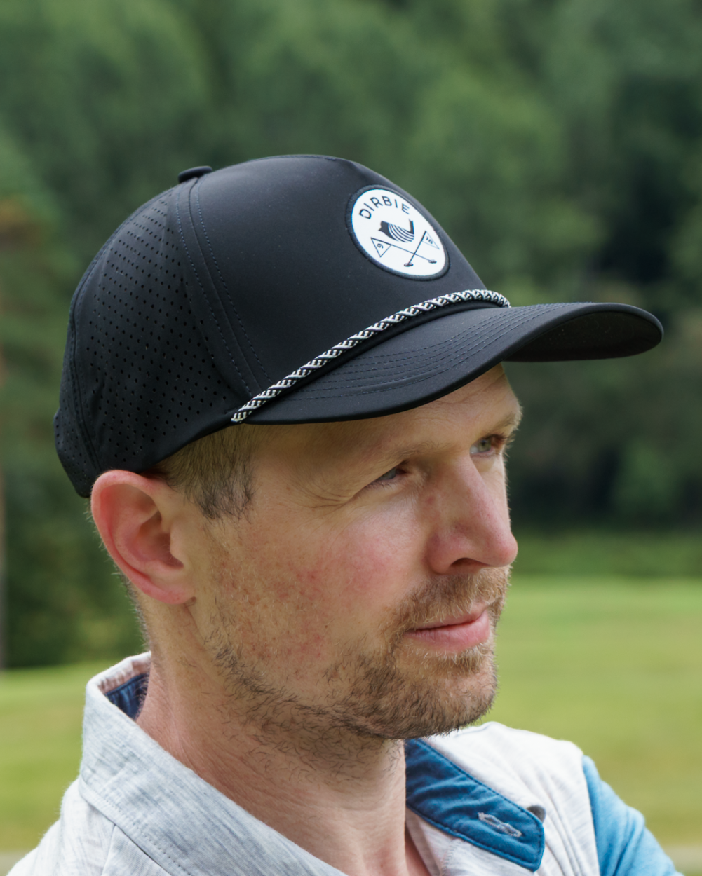 A man wearing a black mesh golf cap with a circular white "Dirbie" logo on the front, photographed outdoors against a blurred green forest background.